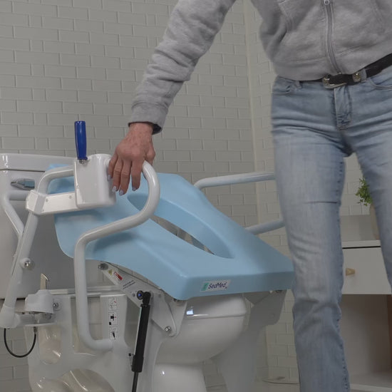 A person in blue jeans is holding the handrail of a SedMed Toilet Lift Assist device, which has a blue seat and side handles for safety.