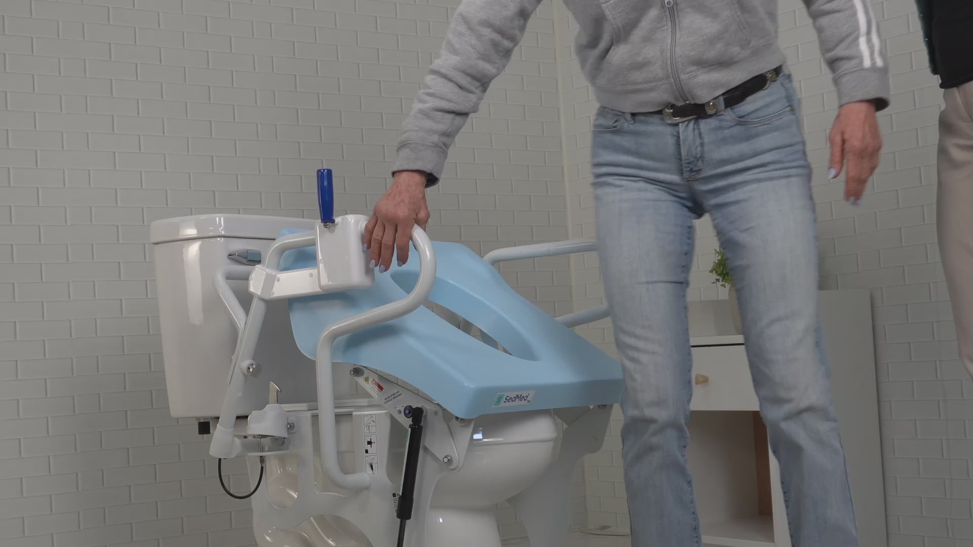 A person in blue jeans is holding the handrail of a SedMed Toilet Lift Assist device, which has a blue seat and side handles for safety.